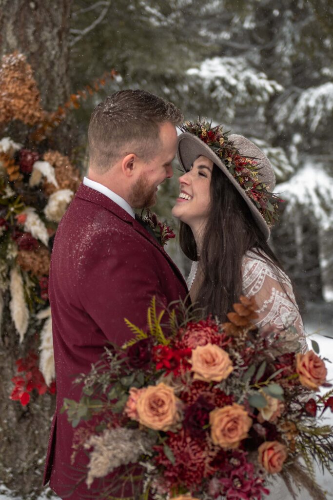 bride in lacey winter dress and flower wool had and groom in maroon suit with red and orange flowers elope in boho wedding